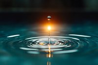 Water droplet creating ripples on calm surface with golden sunlight reflection and dramatic sky background for wellness and meditation concepts.