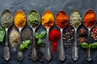Colorful spices and herbs in vintage metal spoons arranged in row on dark slate background, aromatic cooking ingredients for culinary use.