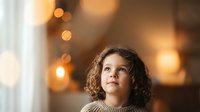 Young mixed-race girl with curly hair looking up thoughtfully in warm indoor lighting with bokeh background for family lifestyle content.