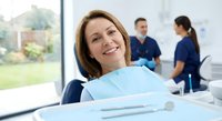 Middle-aged Caucasian woman smiling in dental chair during checkup with medical professionals in background at modern clinic for oral health care.