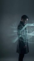 Young man with dark hair and light complexion wearing dark coat surrounded by glowing digital wireframe graphics and data visualization elements against moody gray background.