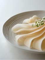 Fresh thin sliced white cheese arranged in elegant waves on ceramic plate, garnished with micro greens, minimalist food styling on light background.