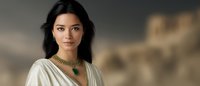 Young Asian woman in white draped dress and ornate gold necklace with emerald stone against blurred ancient architecture background, dramatic portrait lighting.