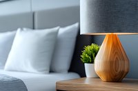 Modern wooden teardrop table lamp with gray shade on nightstand next to white bed pillows and small green plant, contemporary bedroom interior design detail.