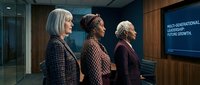 Three mature women of diverse ethnicities stand in a modern office conference room, viewing a digital screen displaying multi-generational leadership and future growth text during a business meeting.