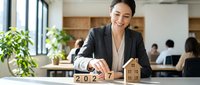 Woman in business attire arranging wooden blocks with 2027 and house symbol at desk in modern office with colleagues and plants in background for real estate planning.