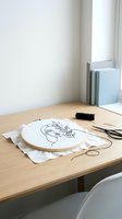 Embroidery hoop with floral line art design on fabric, crafting supplies and scissors on wooden desk in bright modern workspace for DIY projects.