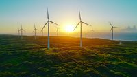 Wind turbines on green hills at golden sunset, aerial view of sustainable renewable energy farm with dramatic sky and sunlight through spinning blades.