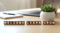 Small succulent plant in white pot on wooden desk with laptop, notebook and wooden blocks spelling welcome learn grow for office decor and workspace motivation.