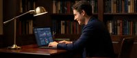 Young professional businessman working on laptop with financial data charts in elegant home office library with warm lighting and bookshelf background.