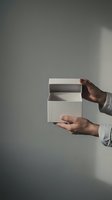Hands opening white cardboard box against gray wall background for product packaging and delivery concepts.