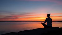 Male silhouette meditating in lotus position on rocky shore during golden sunset over calm water for wellness and mindfulness concepts.