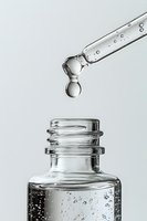 Clear liquid dropper with bubbles suspended above glass bottle containing transparent serum against light background. Macro photography captures water drops.