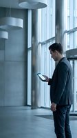 Young professional businessman using tablet computer in modern office building lobby with large windows and contemporary architecture.