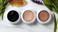 Natural clay face mask products in black and nude tones arranged with honeycomb, lavender flowers and green leaves on white marble background for spa and beauty care.