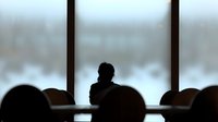 Silhouette of person sitting alone in empty conference room against large window with snowy winter landscape outside, creating moody atmospheric scene.