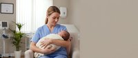 Caucasian female nurse in blue scrubs holding newborn baby in modern hospital room. Healthcare professional providing infant care and support.