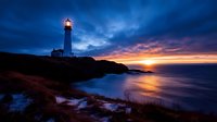 Lighthouse illuminated at dusk with dramatic sunset sky reflecting on ocean water, rocky coastline silhouette creates moody coastal landscape atmosphere.