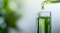 Green liquid drops falling from bottle into glass with bubbles and splashes on blurred natural background, macro photography for wellness and freshness concept.