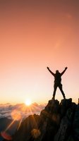 Silhouette of victorious hiker with raised arms standing on mountain peak at sunrise with dramatic orange sky and sun rays breaking through clouds.