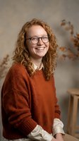 Young Caucasian woman with curly red hair wearing glasses and orange sweater smiling warmly in modern office setting for professional headshot photography.