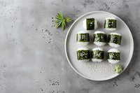 Fresh sushi rolls with nori seaweed and rice served on white ceramic plate against grey concrete background with wasabi and sesame seeds.