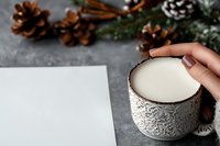 Female hand holding ornate ceramic mug with milk or white drink, blank paper sheet, pine cones and winter decorations on grey concrete background.