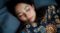 Young Asian woman sleeping peacefully on blue bedding, wearing traditional floral patterned dress. Soft natural lighting creates serene atmosphere for wellness and lifestyle concepts.