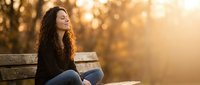 Young woman with curly hair sitting on wooden bench during golden hour sunset, peaceful moment of relaxation and contemplation in autumn park setting.