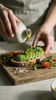Woman drizzling olive oil on avocado toast with cherry tomatoes and microgreens on rustic wooden cutting board for healthy breakfast preparation.