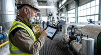 Senior Caucasian male industrial engineer in safety vest using digital tablet for equipment monitoring in modern manufacturing facility.