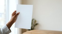 Close-up of a person's hand holding a blank white sheet of paper against a neutral background with natural light, suitable for mockups and presentations.