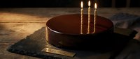 Dark chocolate birthday cake with three lit golden candles on rustic wooden table, milestone 30 brass plaque, moody dramatic lighting for celebration.