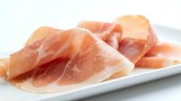 Thinly sliced prosciutto ham arranged on white ceramic plate, ready to serve as gourmet appetizer or charcuterie board ingredient for culinary presentations.