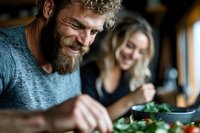 Mature bearded man with curly hair enjoying healthy meal at restaurant with blonde woman companion smiling in background during lunch date.