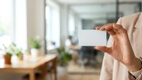 Businesswoman holding blank white card in modern office workspace with natural light and green plants, suitable for corporate branding and business communication concepts.