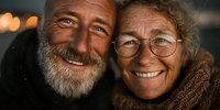 Happy senior couple smiling together outdoors with warm bokeh lights in background. Elderly man with beard and woman with glasses showing love and joy.