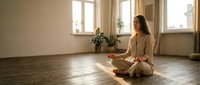 Young Caucasian woman meditating in lotus pose on wooden floor in sunlit studio, practicing mindfulness and inner peace for wellness content.