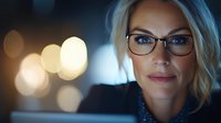 Professional blonde businesswoman with glasses working late at office computer with bokeh city lights background for corporate presentations.
