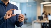 Man in navy button-up shirt holding blank business card in modern office environment with colleagues in background, suitable for corporate branding and professional networking materials.