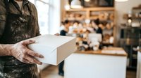 Male worker with weathered hands holding white cardboard box in bakery or café setting with blurred background, suitable for small business, delivery, and retail concepts.