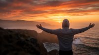 Unrecognizable person with arms outstretched standing on coastal cliff at golden sunset, celebrating freedom and achievement with ocean waves below.