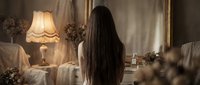 Young woman with long dark hair in elegant bedroom interior with vintage lamp and mirror creating atmospheric mood for lifestyle photography.