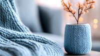 Cozy knitted blue vase with dried flowers and warm blanket creating hygge atmosphere. Soft bokeh lights in background add warmth to minimalist home decor.