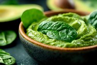 Fresh avocado guacamole dip garnished with baby spinach leaves in rustic ceramic bowl on dark background, close up view of healthy vegan appetizer.