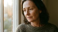 Mature woman with long dark hair and fair complexion gazing thoughtfully out window, wearing knit sweater, natural indoor lighting, contemplative mood suitable for lifestyle, wellness, mental health, aging, and editorial content.