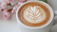 Creamy latte art with delicate rosetta pattern in white ceramic cup against soft pink floral background, selective focus on coffee foam design.