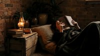Man relaxing on couch in cozy living room with warm lighting, reading book near lamp and brick wall for home decor and lifestyle content.