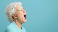 Senior Caucasian woman with white hair yawning against turquoise background, expressing tiredness and natural human behavior in minimalist studio setting.