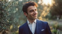 Young Mediterranean man in navy pinstripe suit smiling confidently outdoors with golden sunset light and olive trees creating natural bokeh background.